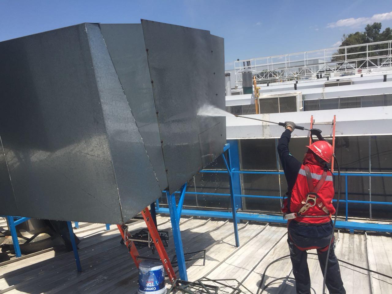 Trabajador limpiando estructura metálica con manguera de alta presión en un techo, usando casco y arnés de seguridad.