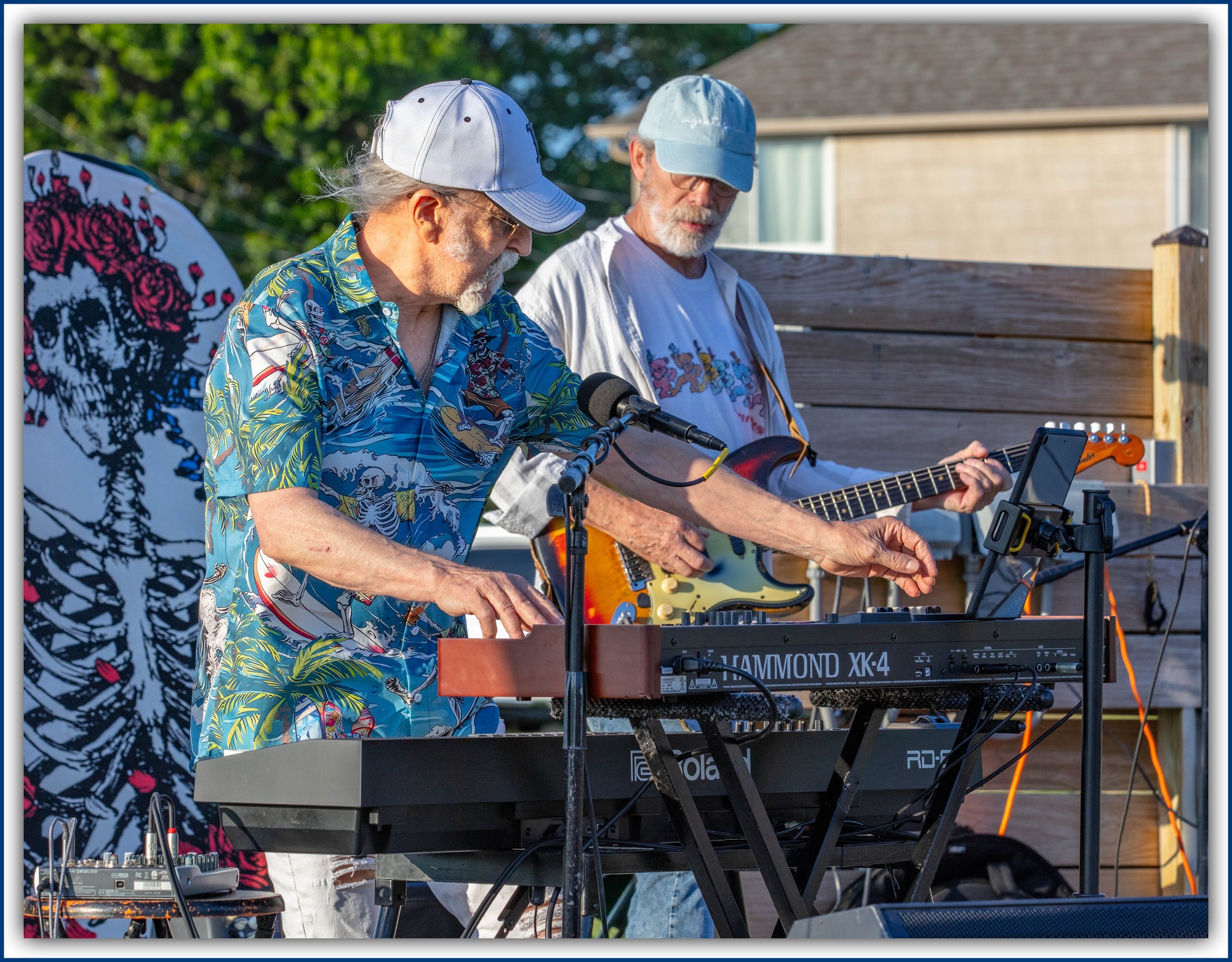 Man with a beard and glasses playing electric guitar outdoors, wearing a t-shirt with a skull logo and a cap.