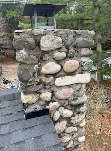 Stone chimney with metal cap on rooftop, surrounded by woodland area