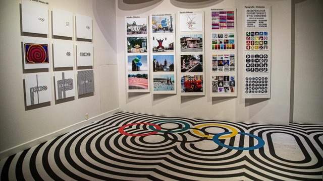 Exposición de afiches y logotipos olímpicos en una galería, mostrando obras gráficas en las paredes y un diseño de aros olímpicos en el suelo.