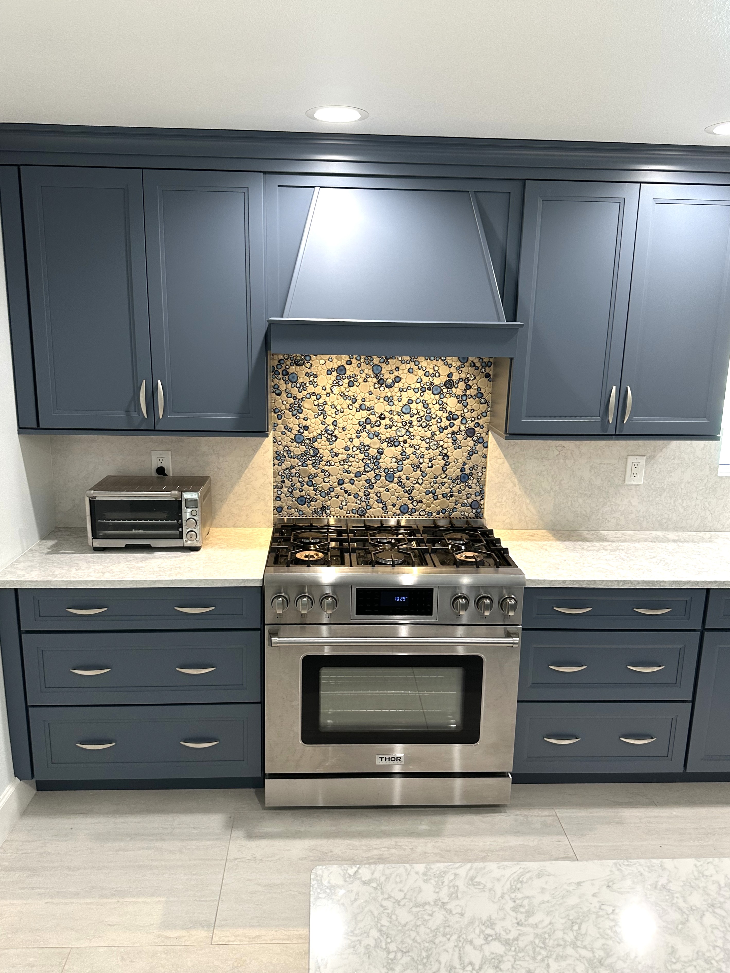 The expertly tiled backsplash behind the stove enhances the beachy blue hood and cabinetry, creating a stunning focal point in the kitchen.