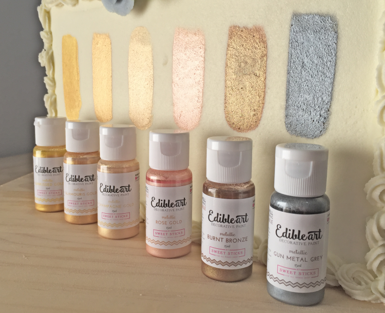 Bottles of Edibleart decorative paint in metallic shades, with color swatches on a cream-colored surface, showcasing shades like rose gold and gun metal grey.