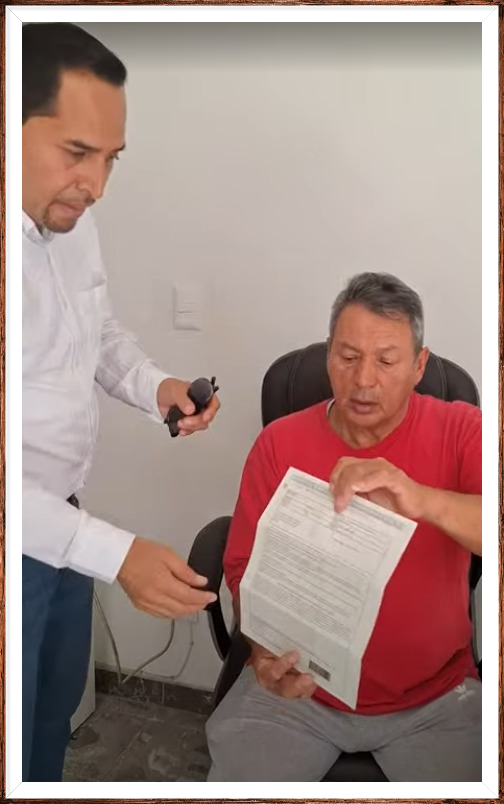 Dos personas conversando en una oficina, una mujer sostiene un dispositivo y un hombre con camisa azul lleva carpetas amarillas, estante con libros y un globo terráqueo detrás.