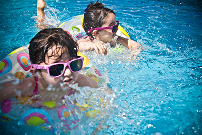 Two children wearing sunglasses playing in a swimming pool with colorful inflatable rings on a sunny day.