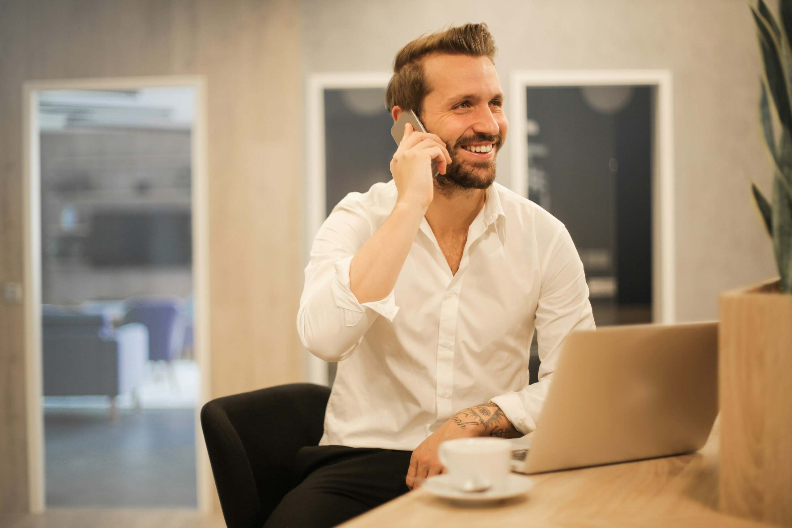 Hombre sonriente con camisa blanca hablando por teléfono en una oficina moderna, con una computadora portátil y una taza de café en la mesa.