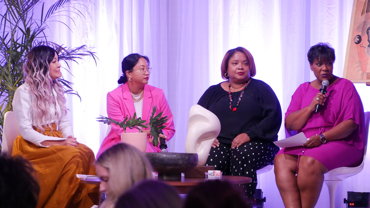 Smiling woman in checkered dress with curly hair, standing indoors in front of a plant and framed photos.