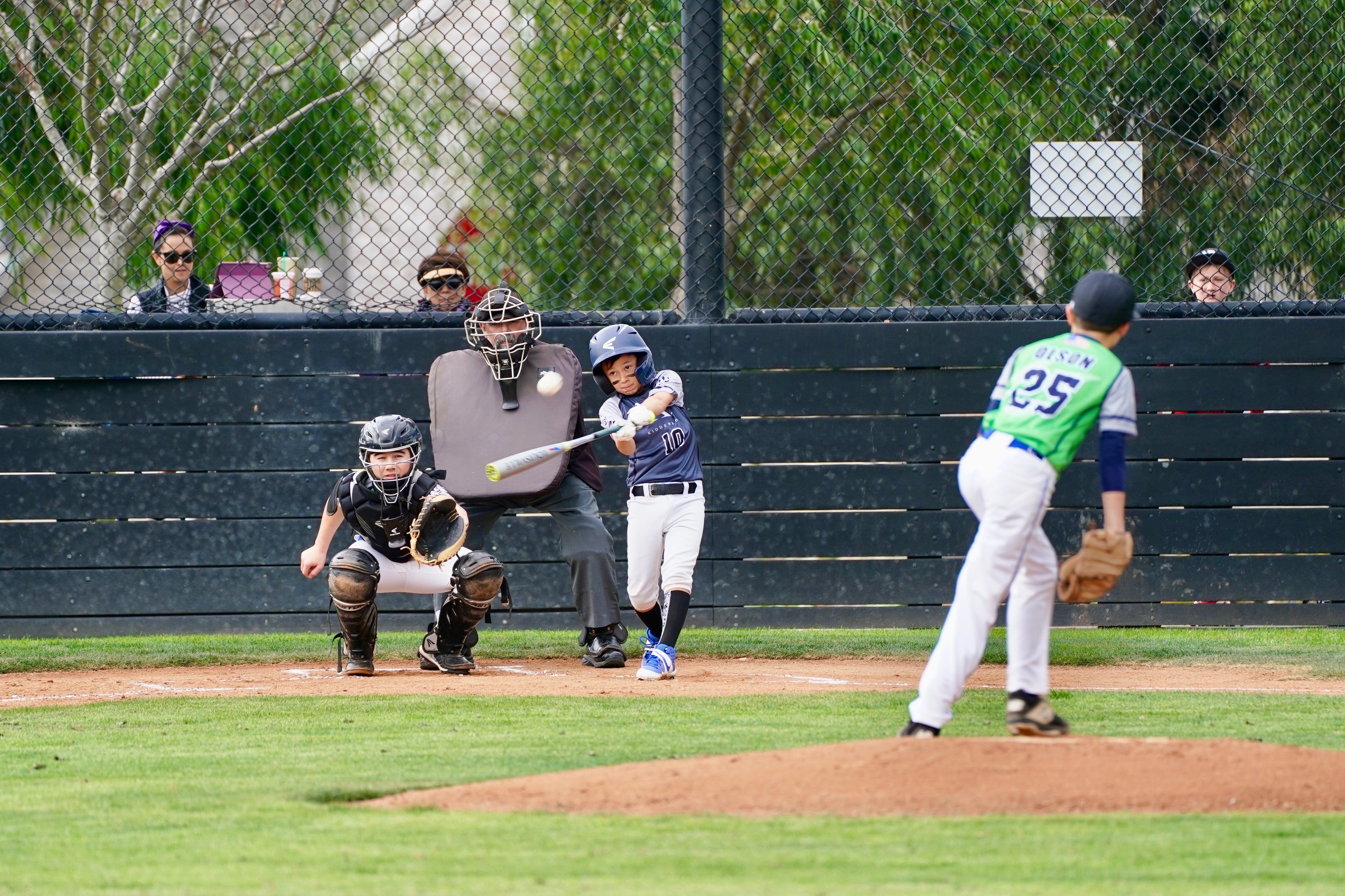 Youth baseball player in blue uniform hitting the ball during a game with catcher, umpire, and pitcher visible.