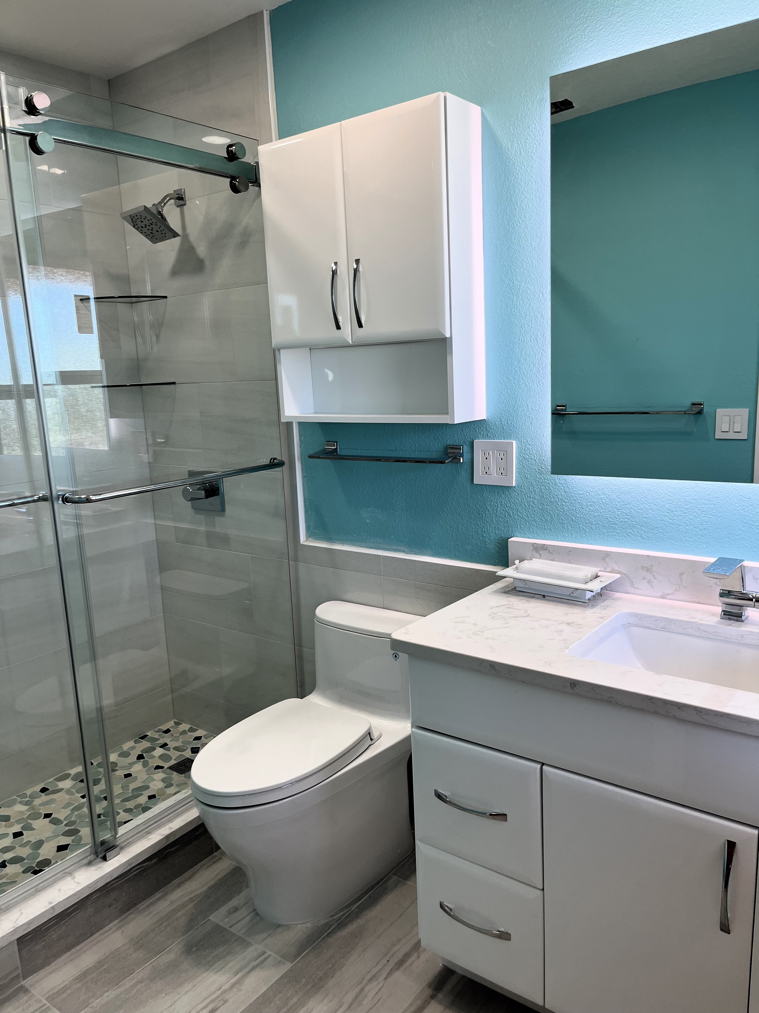 Spacious tiled shower with pebble floor and heavy clear glass by-passing barn doors with dual corner shelves and a large showerhead.  Modern slab front white gloss vanity with matching toilet topper and chrome handles.