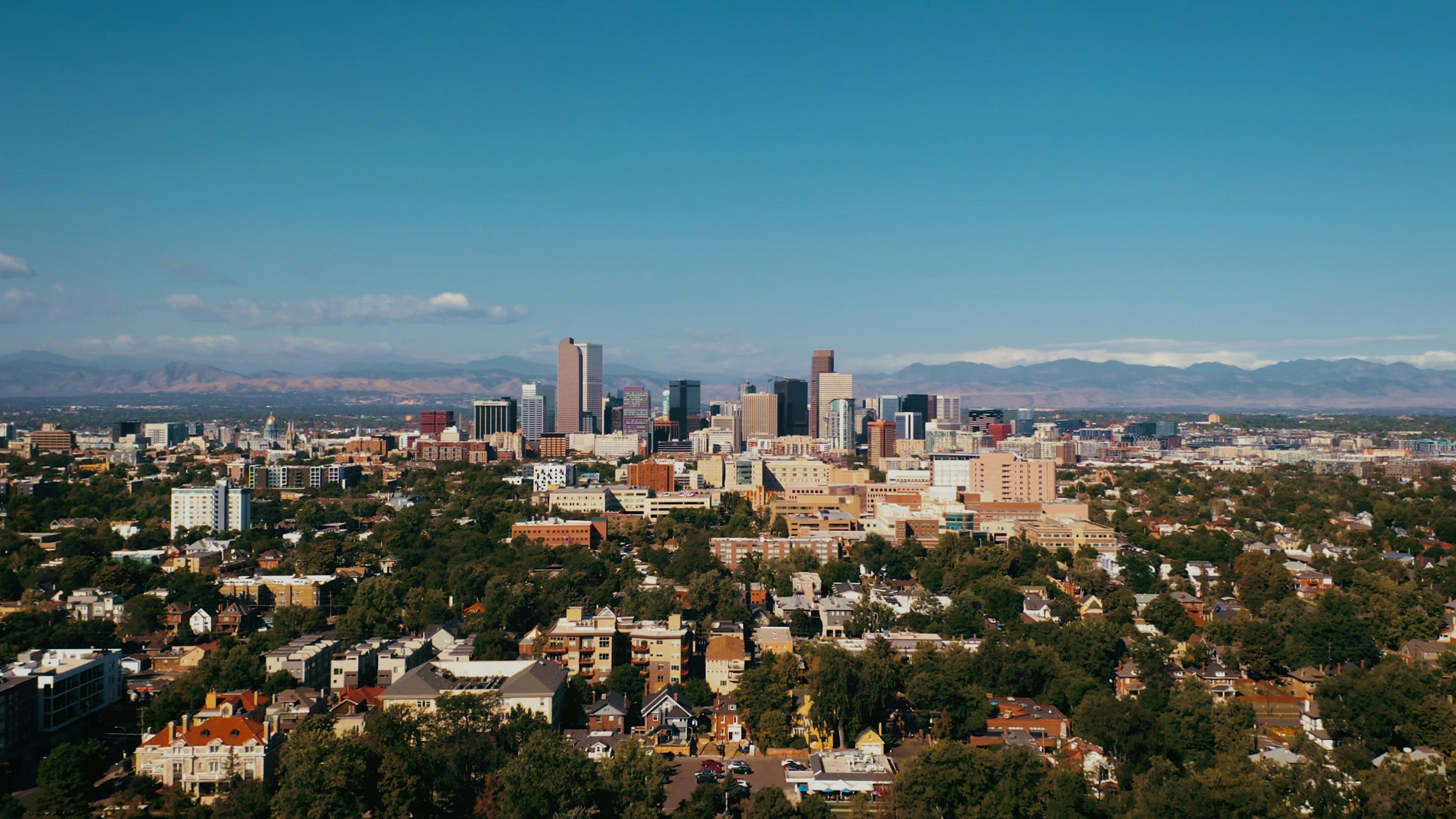 Aerial view of Denver skyline with modern skyscrapers, lush green trees, residential areas, and distant mountains under a clear blue sky.