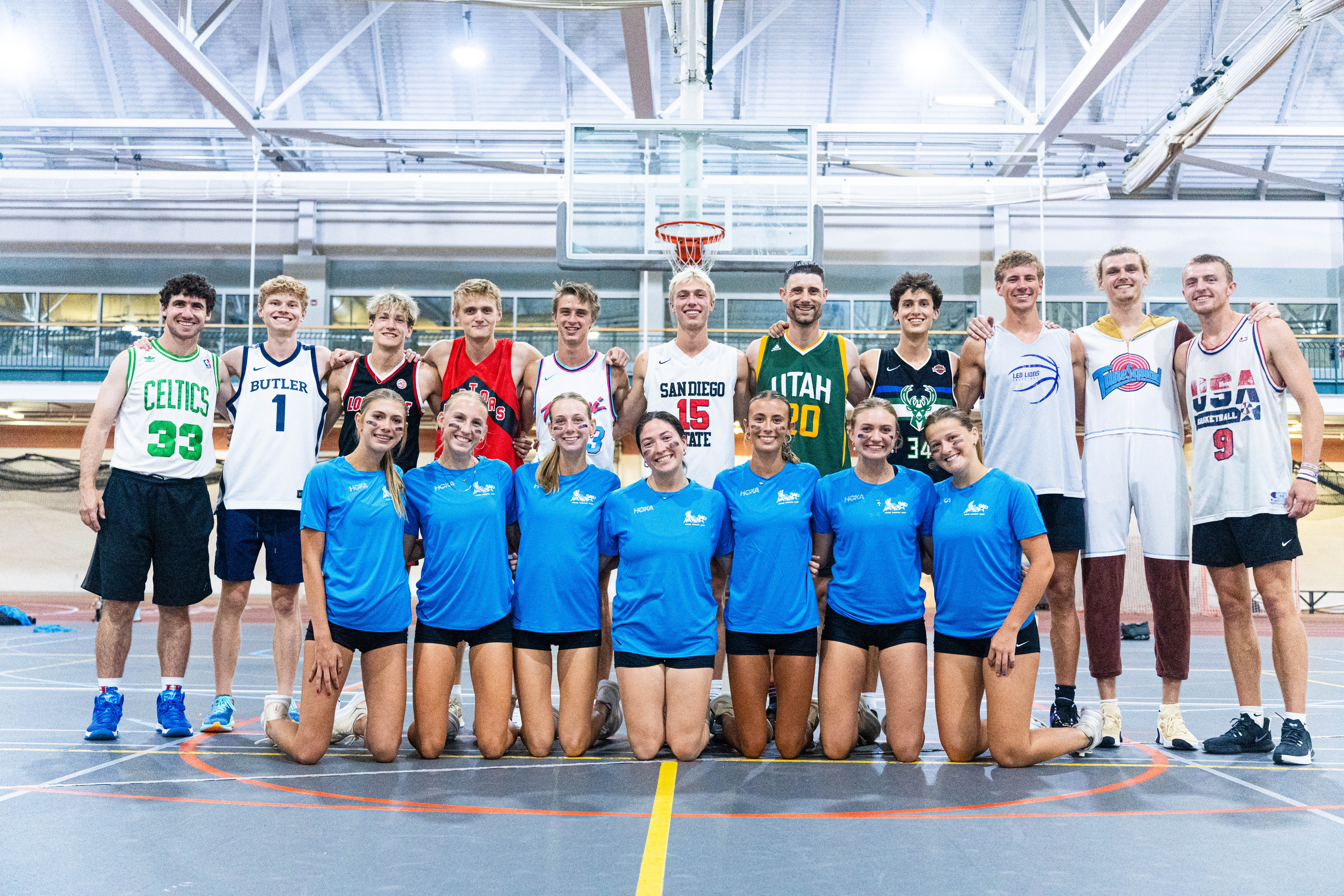 Group of young athletes posing in a basketball court, wearing assorted team jerseys and blue shirts, under a brightly lit gymnasium setting.