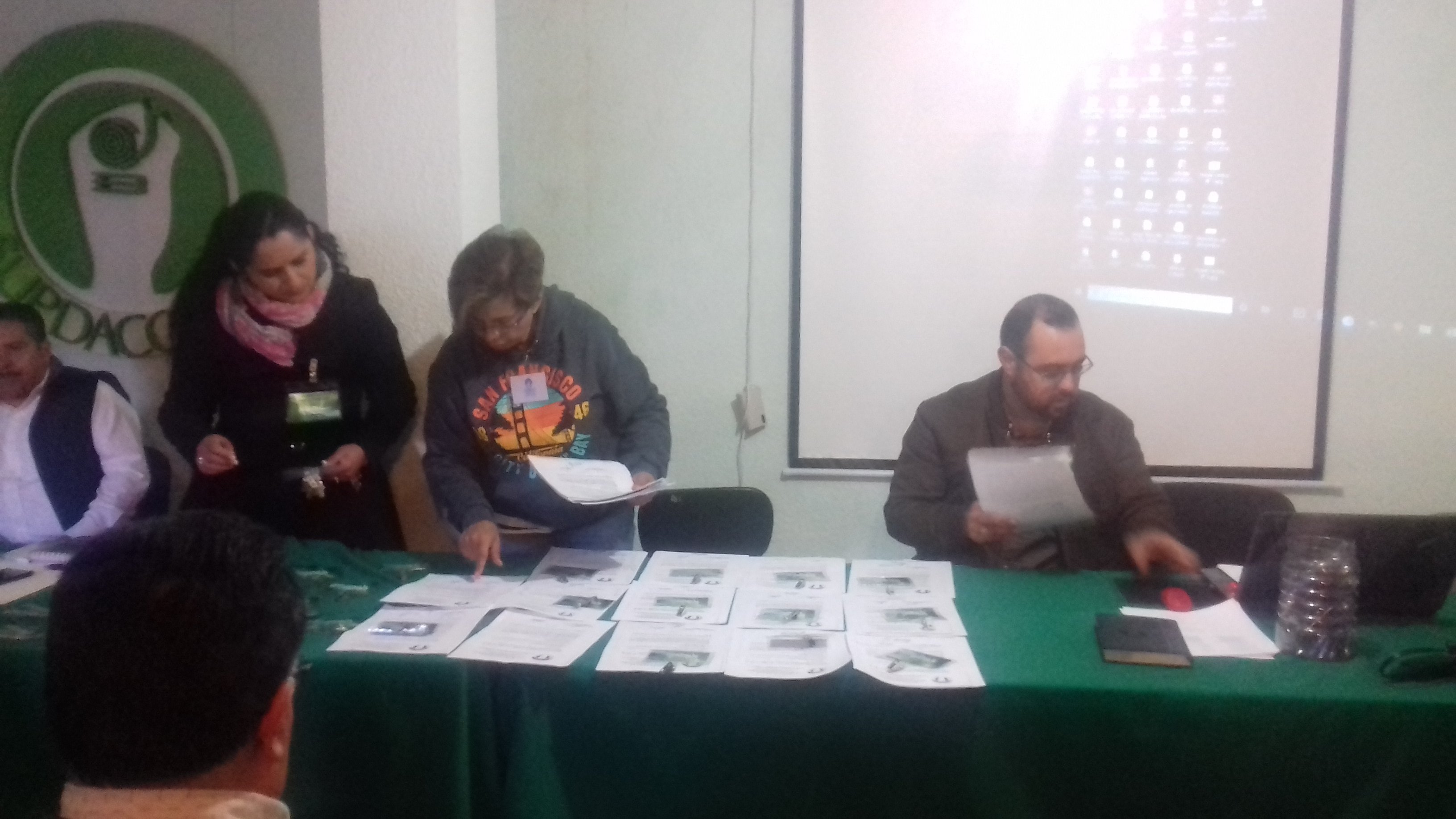 Personas revisando documentos en una mesa verde durante una reunión, con proyector al fondo.