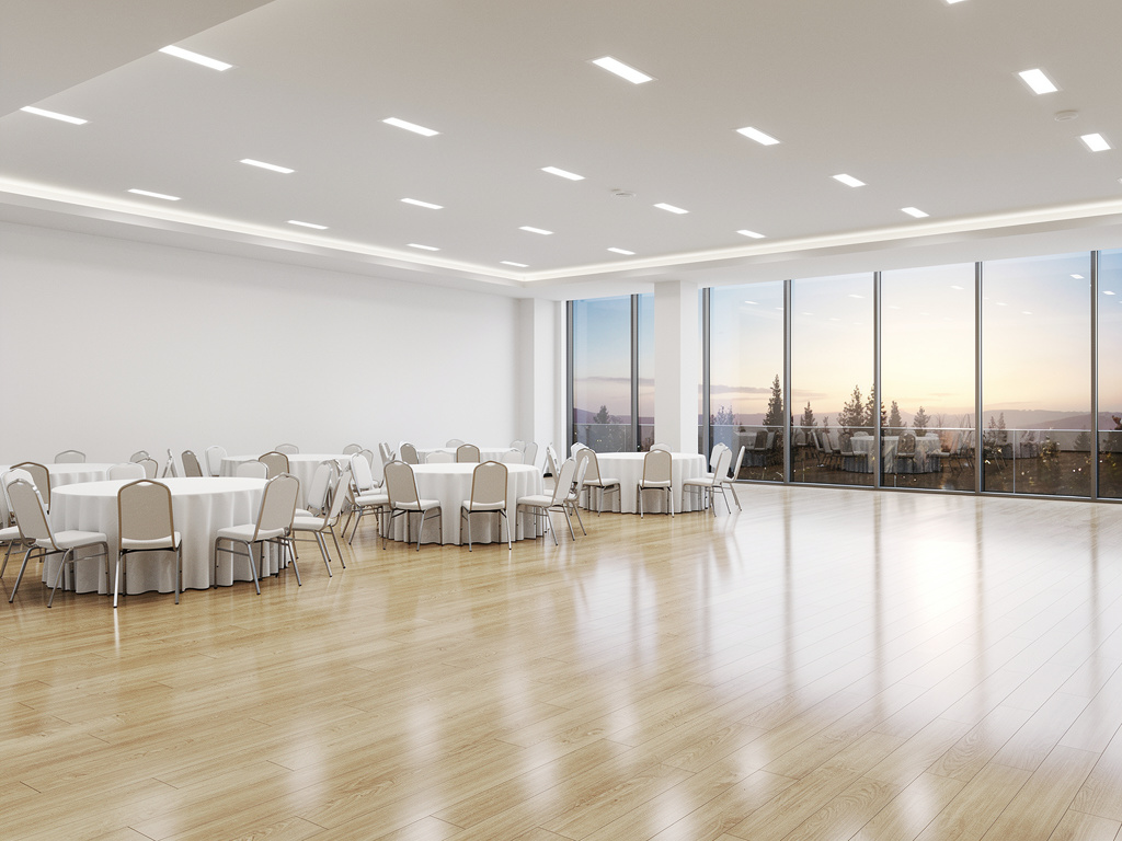 Salón de eventos moderno con mesas redondas, sillas blancas, suelo de madera y grandes ventanales con vista al atardecer
