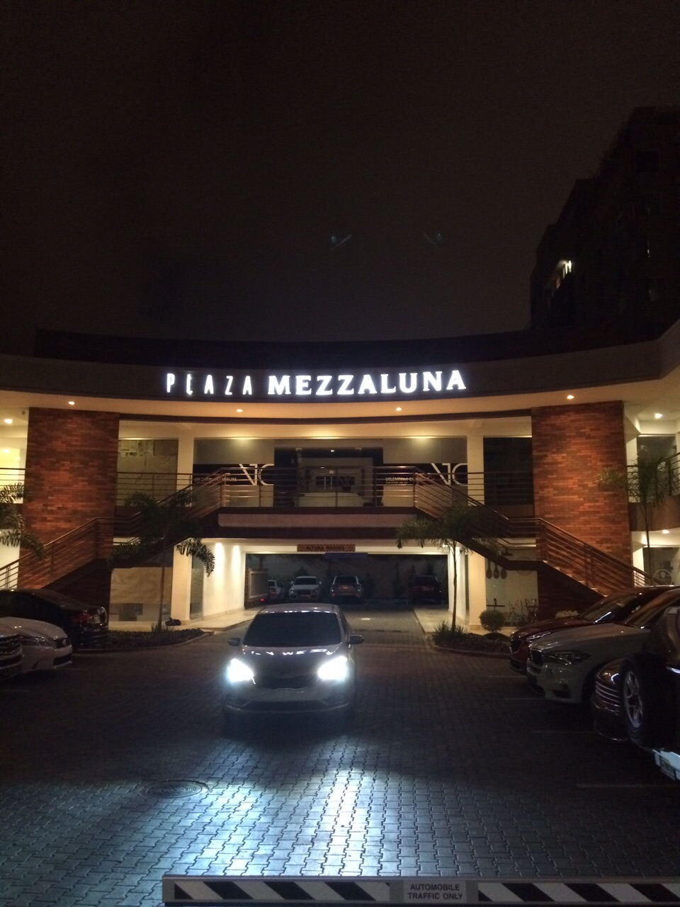 Entrada nocturna de Plaza Mezzaluna iluminada con autos estacionados y vehículo con faros encendidos