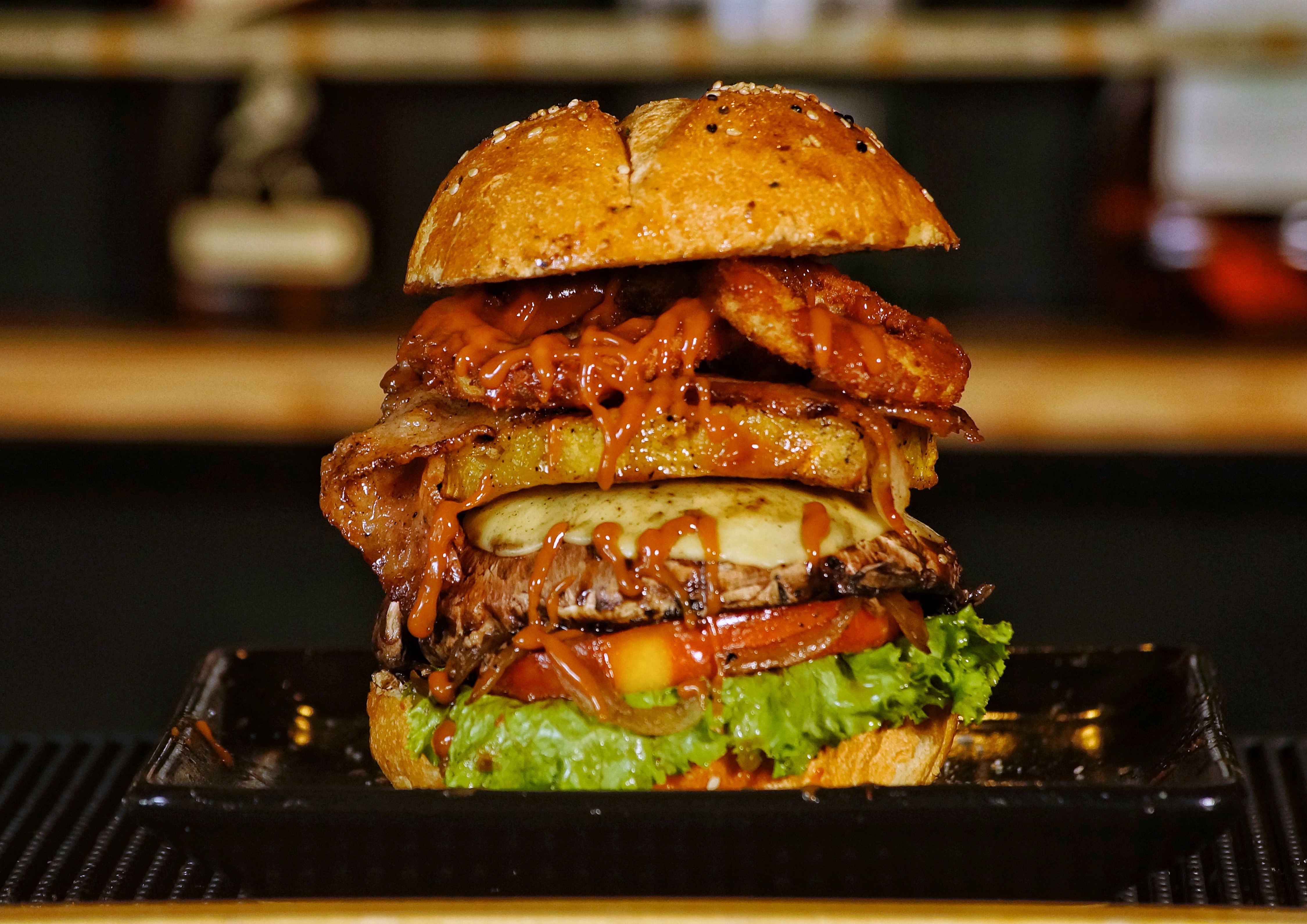 Close-up of a gourmet burger with lettuce, tomato, cheese, meat patty, bacon, onion rings, and sauce on a sesame seed bun on a dark plate.