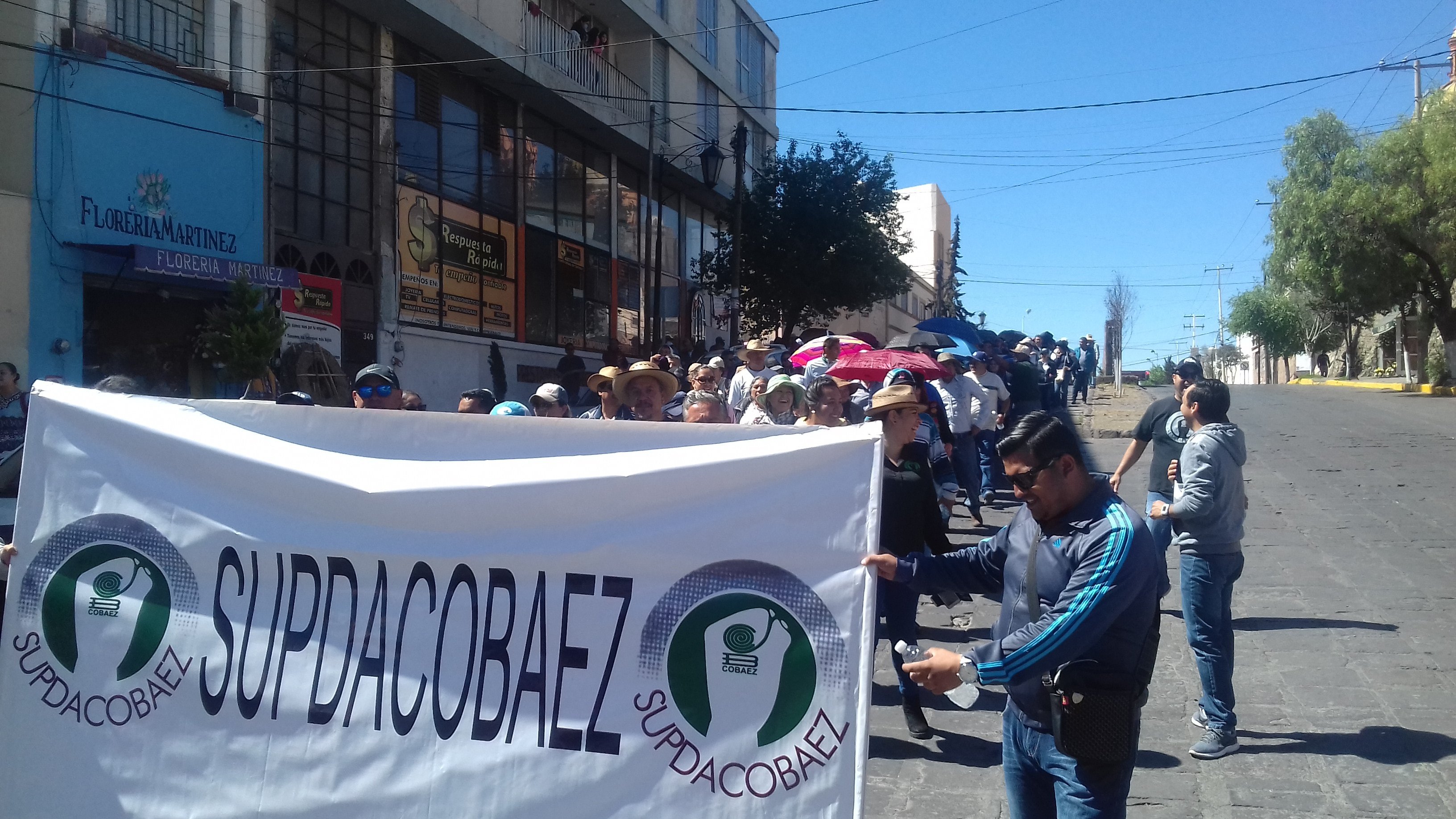 Manifestación en la calle con personas sosteniendo pancarta de SUPDACOBAEZ, algunos protegiéndose del sol con sombreros y paraguas.