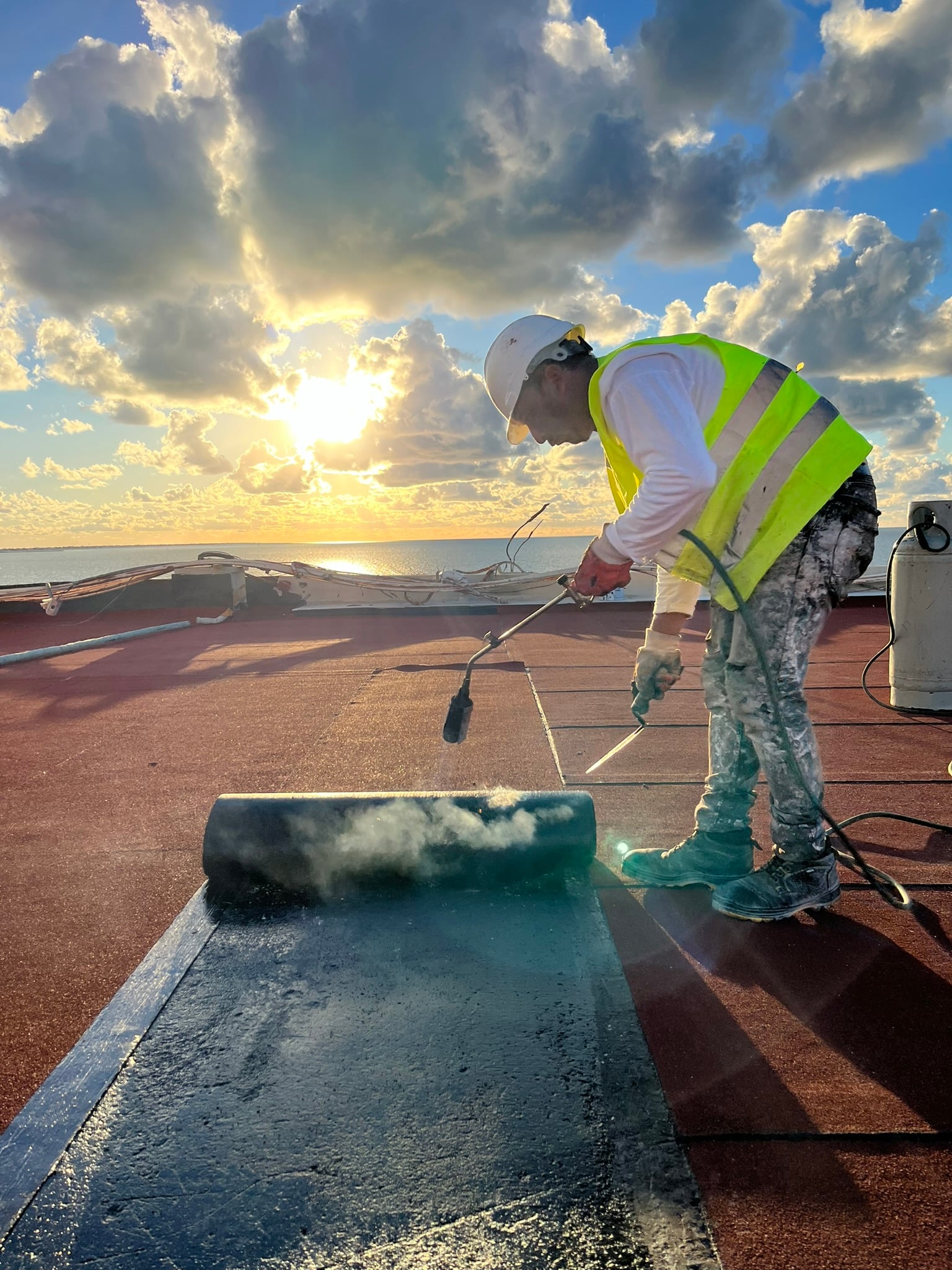 Trabajador en el techo con chaleco reflectante, usando soplete para aplicar material impermeabilizante en la puesta de sol.
