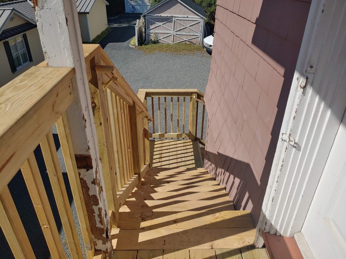 Wooden outdoor stairs with railings leading down to a gravel driveway adjacent to a house wall.