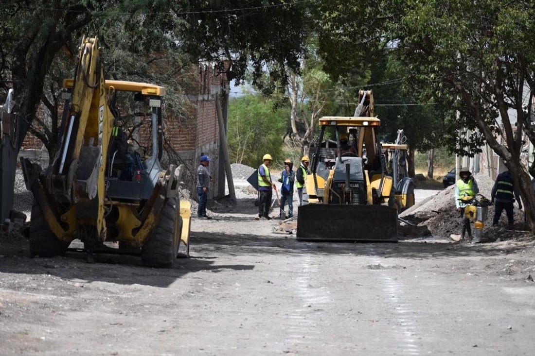 Maquinaria pesada y trabajadores de construcción en una obra vial al aire libre rodeados de árboles.