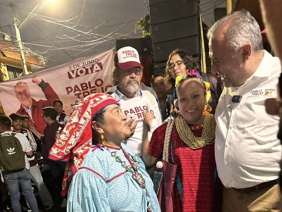 Mujer sonriente sentada en una oficina con fondo decorativo, frente a una mesa de madera y sosteniendo una tableta.