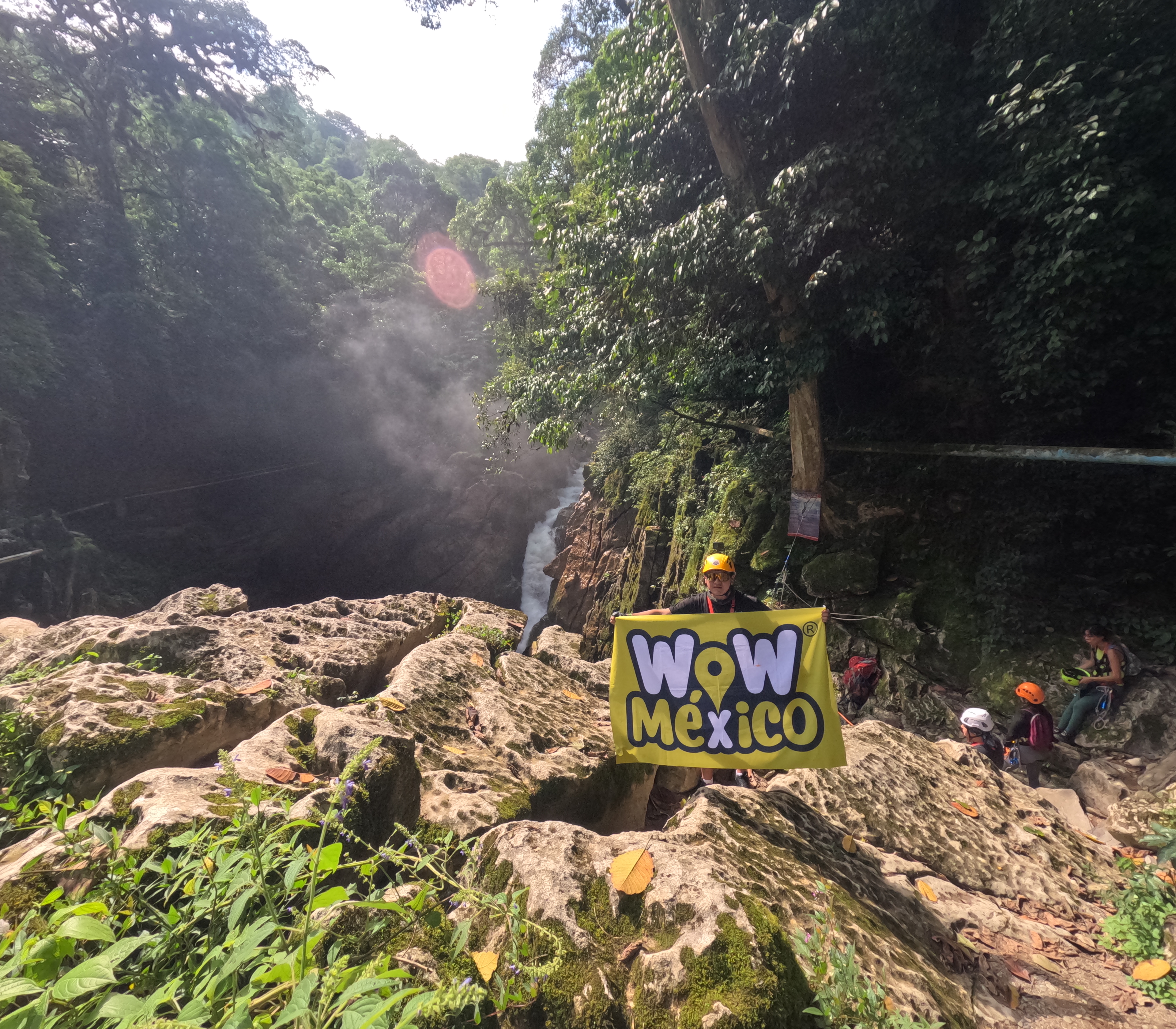 Persona sosteniendo un cartel que dice 'Wow México' en un entorno de selva frondosa con cascada al fondo. Persona sosteniendo un cartel que dice 'Wow México' en un entorno de selva frondosa con cascada al fondo.