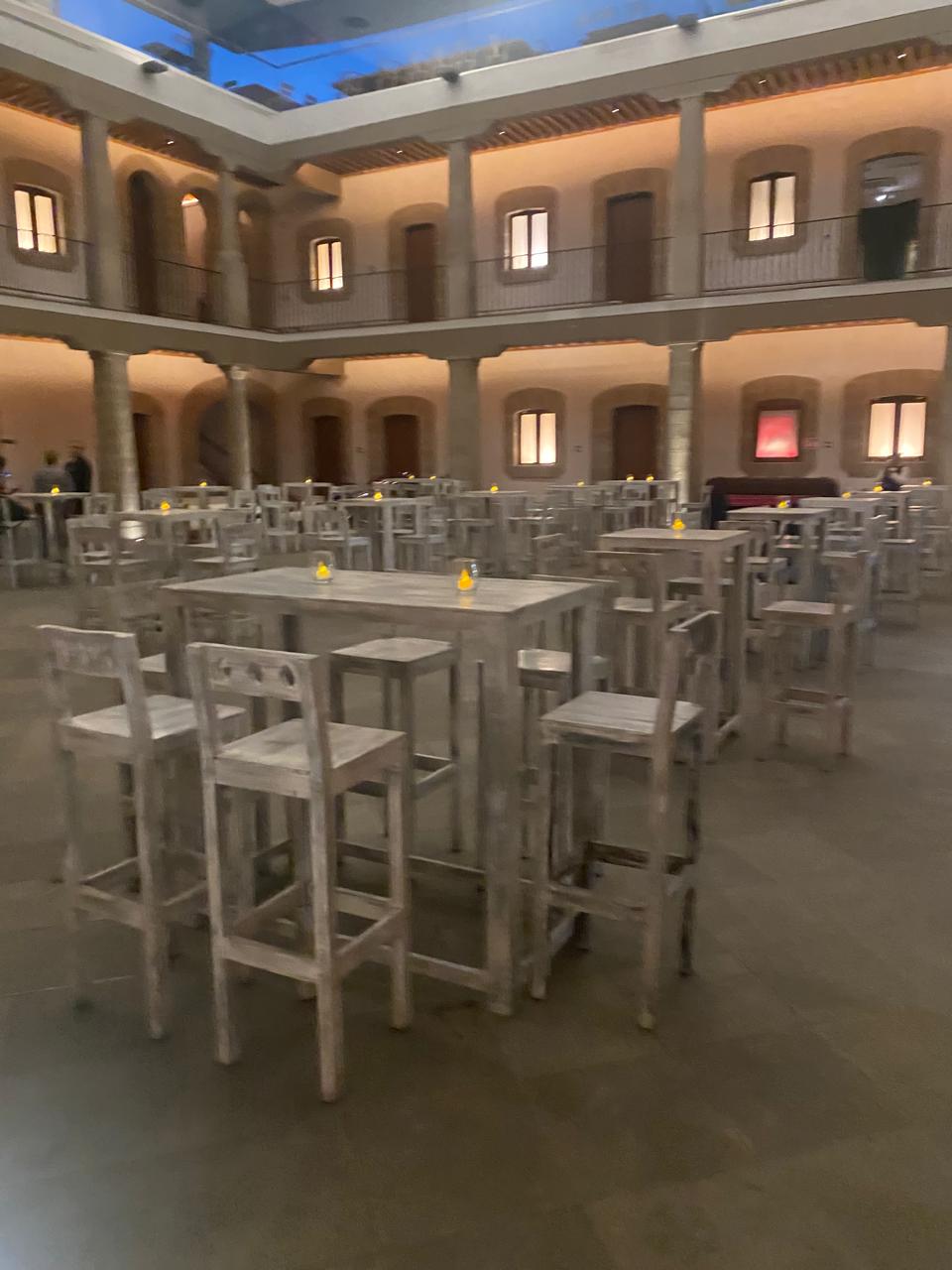 Interior de un edificio con mesas y sillas altas de madera, rodeadas de columnas y balcones iluminados. Interior de un edificio con mesas y sillas altas de madera, rodeadas de columnas y balcones iluminados.