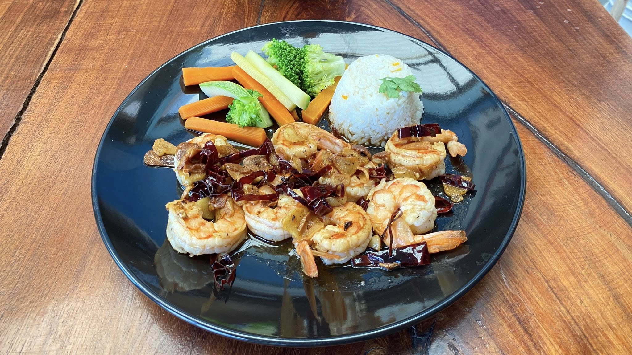 Plato de camarones al ajillo con arroz blanco y verduras al vapor en plato negro sobre mesa de madera. Plato de camarones al ajillo con arroz blanco y verduras al vapor en plato negro sobre mesa de madera.