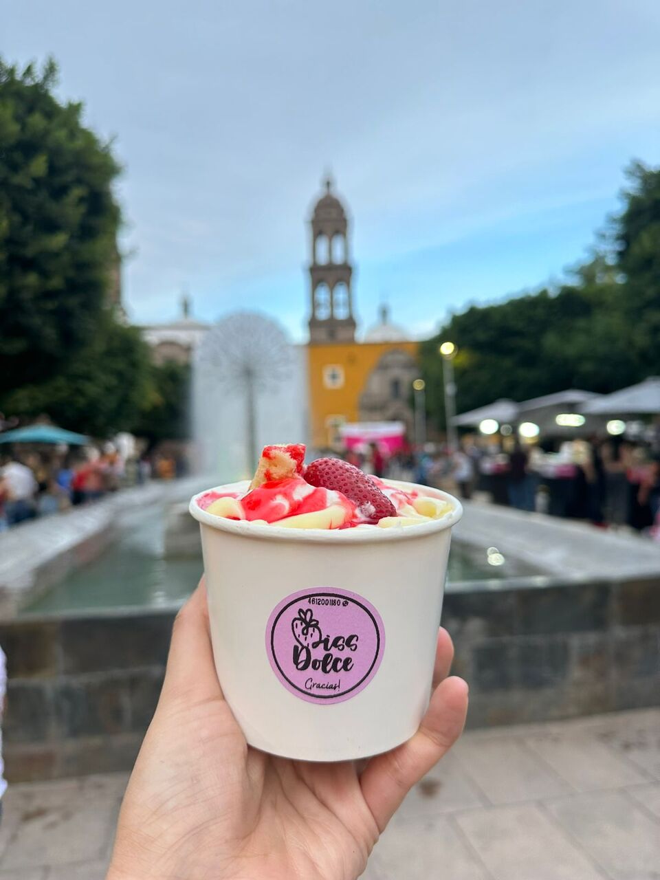 Persona sosteniendo un helado con fresas y jarabe, fondo de plaza con fuente y edificio histórico.