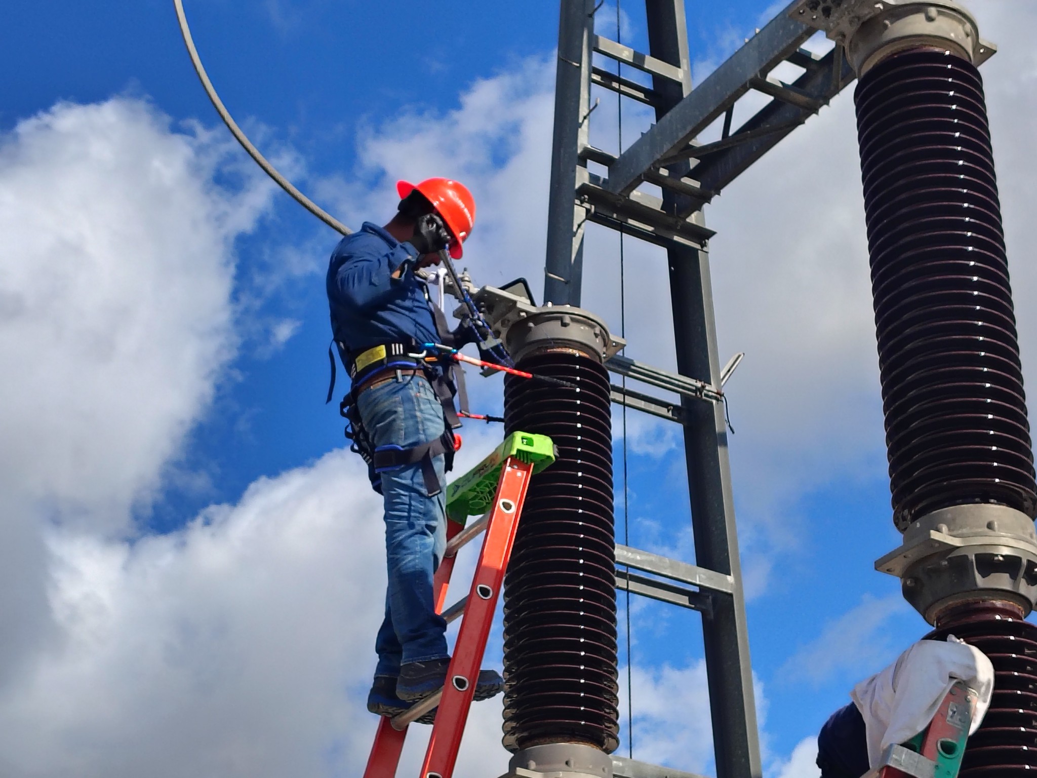 Dos trabajadores con equipo de seguridad inspeccionan un transformador eléctrico verde en una subestación al aire libre bajo un cielo despejado.