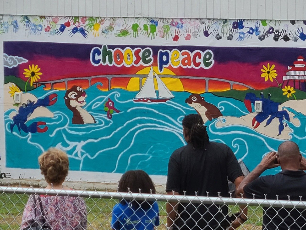 Colorful mural featuring otters, crabs, a sailboat, and the words 'choose peace', viewed by people from behind a fence.