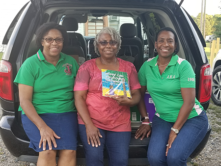 Three women sitting in an open car trunk, smiling and holding a colorful book.