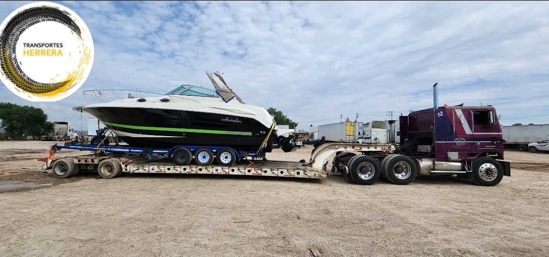 Camión transportando un bote en un remolque de plataforma abierta en un área de carga, con el logotipo de 'Transportes Herrera' en la esquina.