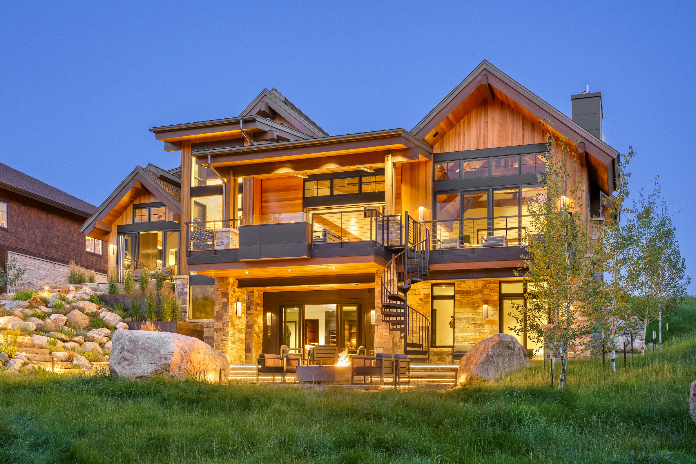 Luxury modern mountain home with large windows, spiral staircase, and patio with firepit at twilight.