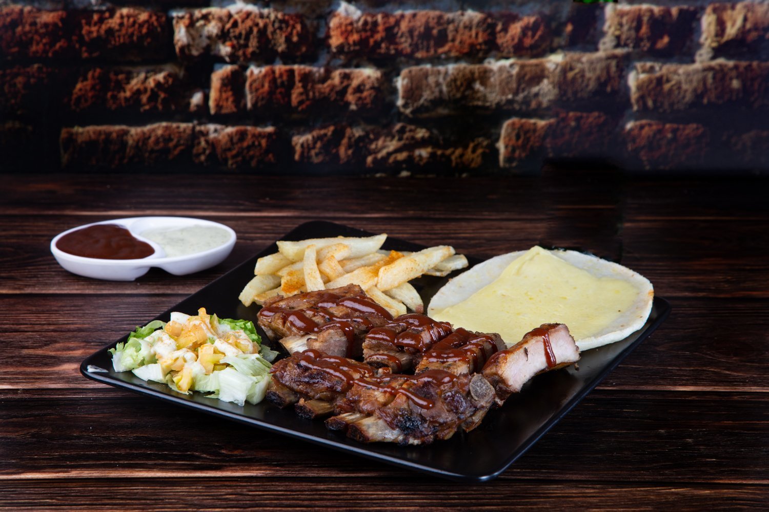 Costillas asadas con salsa BBQ, acompañadas de papas fritas, ensalada y tortilla en un plato negro sobre mesa de madera.