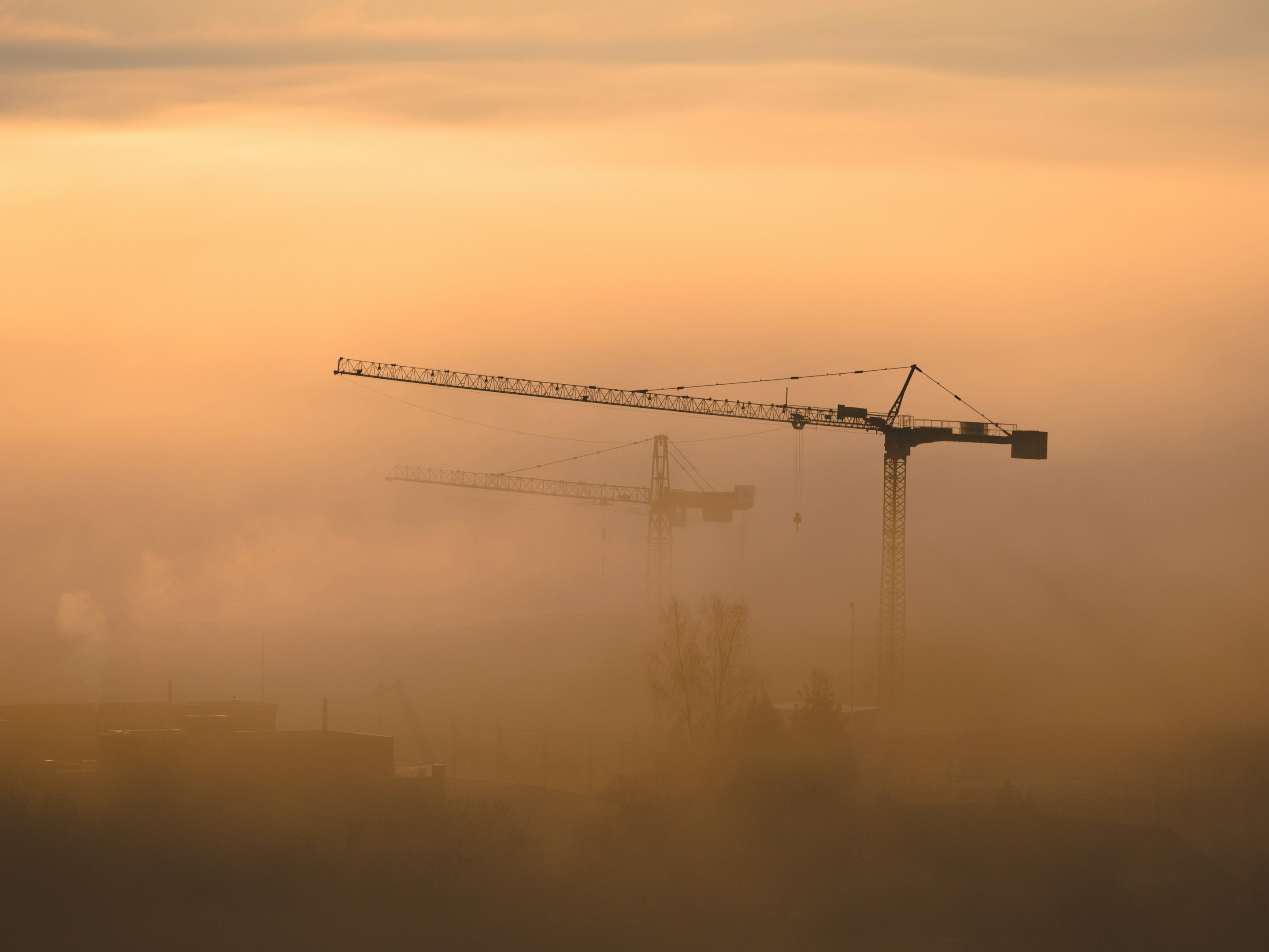 Cranes silhouetted against a hazy golden sunset on a construction site.