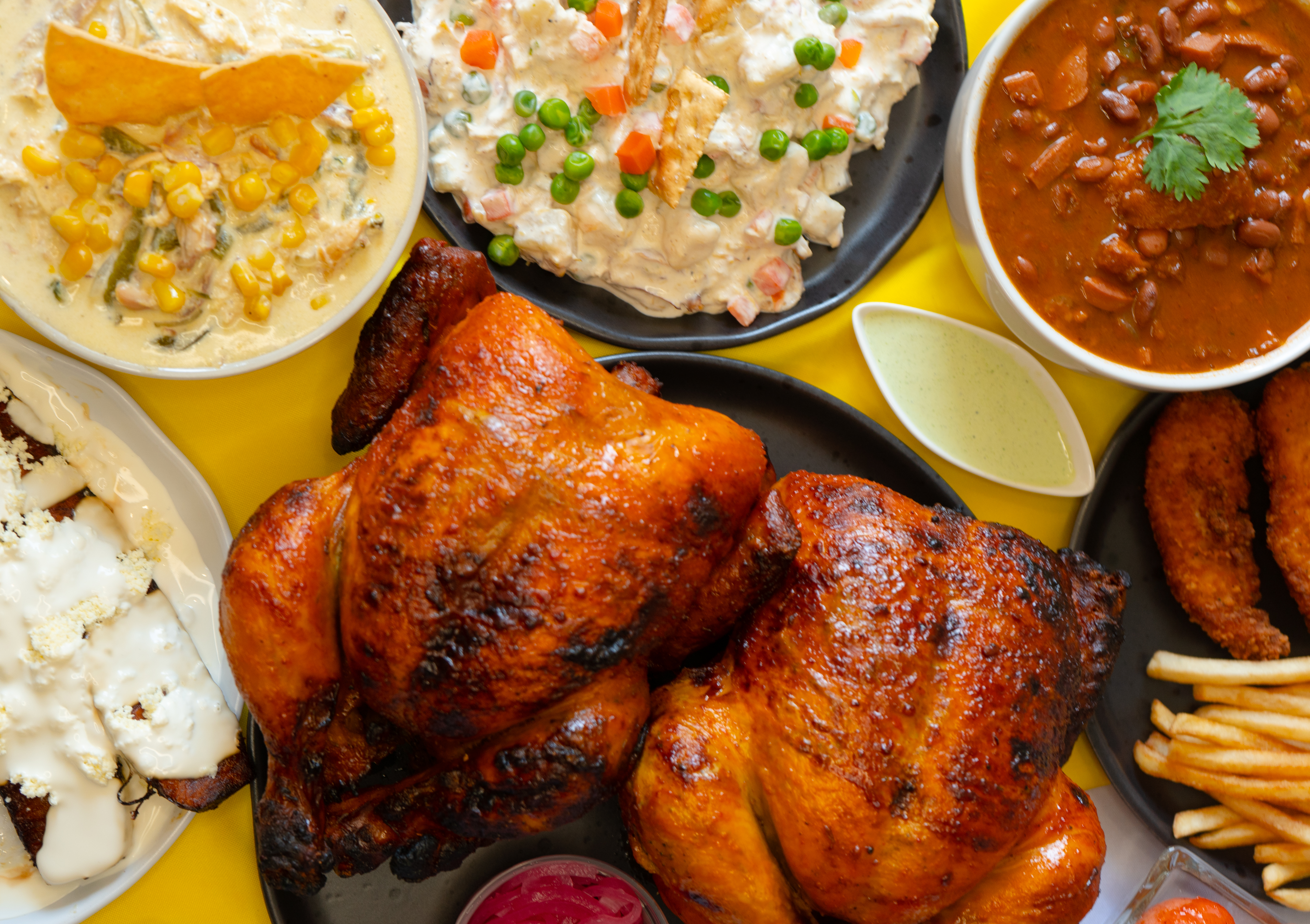 Pollo asado con papas fritas, salsas cremosas, frijoles, maíz, verduras y totopos en mesa amarilla
