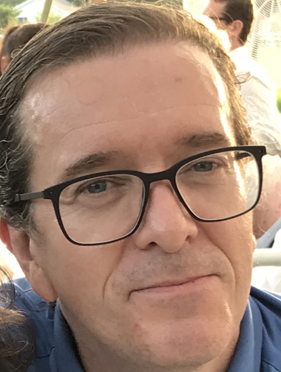 Woman with long hair and glasses smiling in an office with large windows and cityscape view.