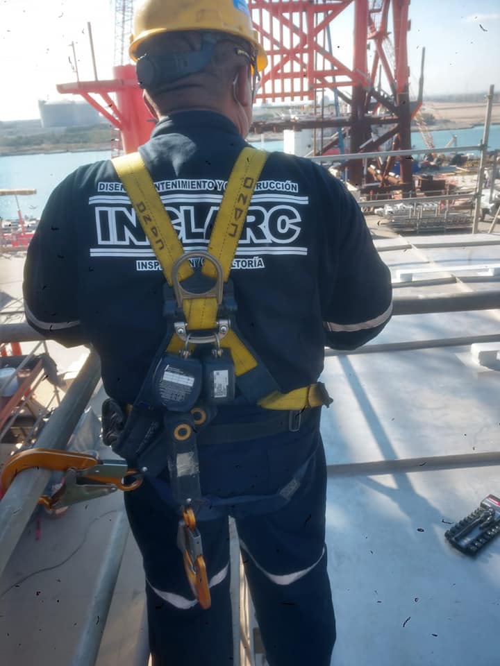 Trabajador de construcción con arnés y casco en un sitio industrial junto a una estructura metálica roja.