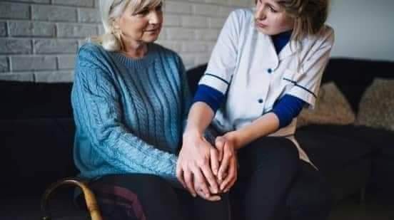Enfermera sentada junto a mujer mayor con bastón, dándole apoyo y atención en sala de estar.