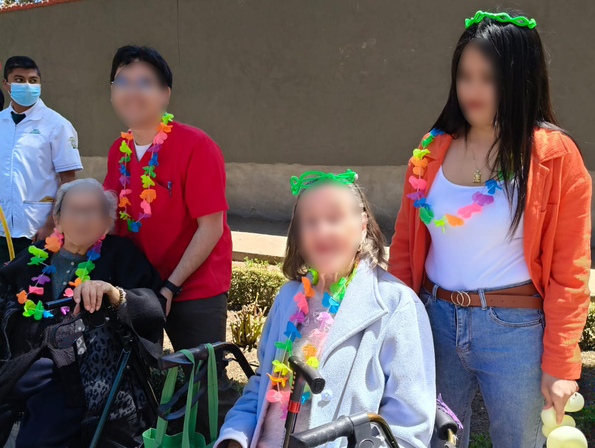 Smiling caregiver with elderly women in wheelchairs holding colorful paper pom-poms at a senior care home