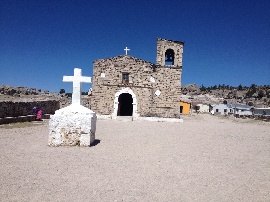Iglesia de piedra con cruz blanca en paisaje rural y cielo azul claro.