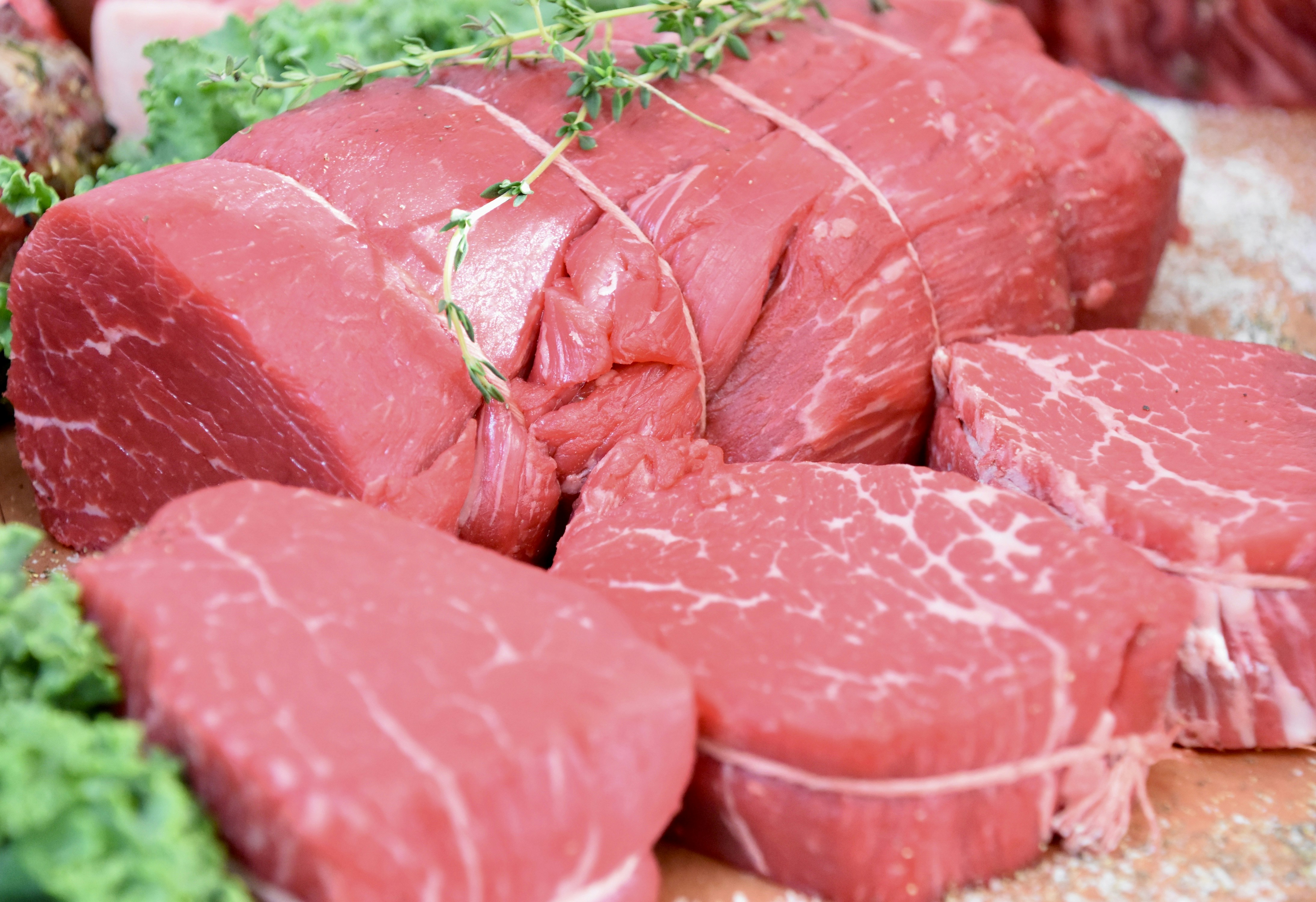 Varios cortes de carne fresca expuestos en una tabla de madera, rodeados de hierbas verdes y tomates en el fondo de una cocina moderna.