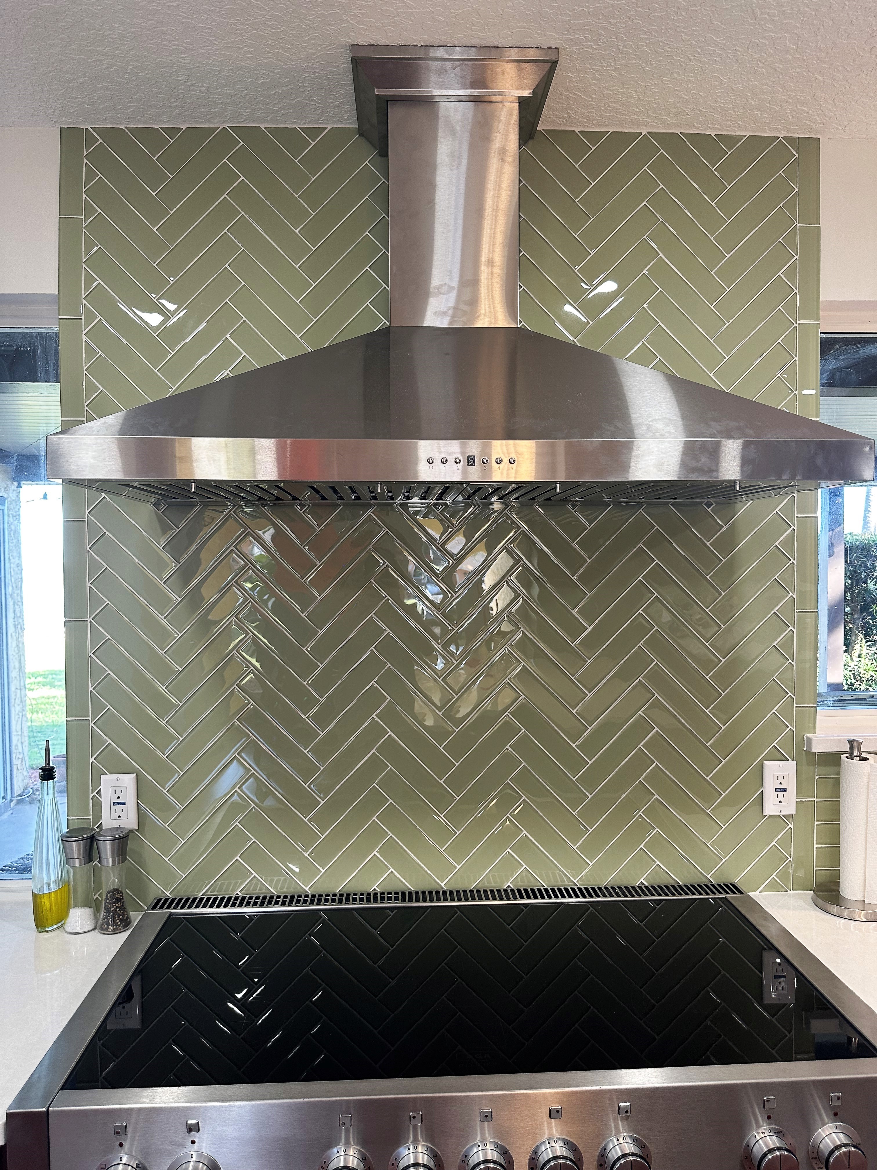 Glossy green backsplash set in a herringbone pattern behind the gourmet oven and vent hood adds an elegant pop of color.