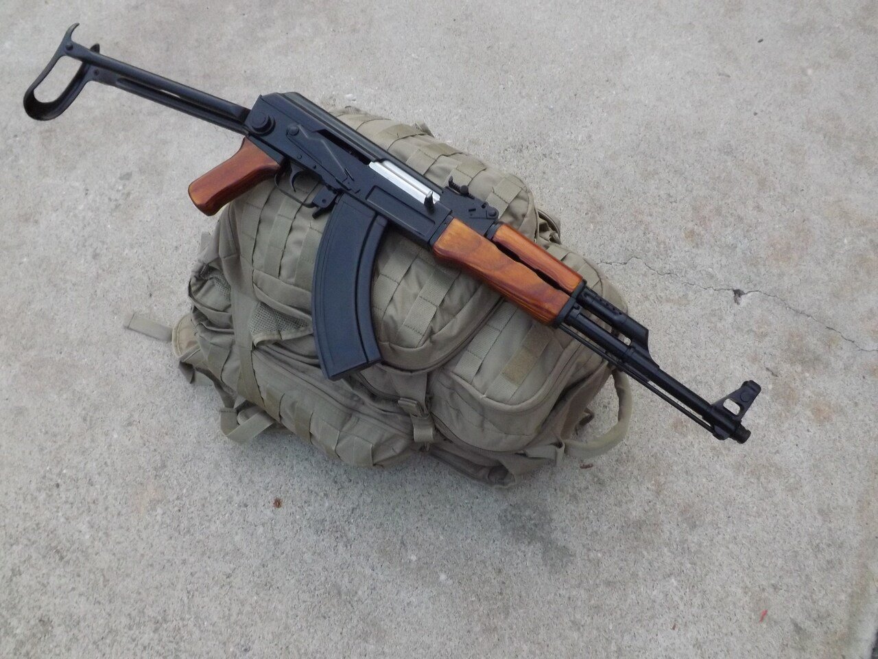Assault rifle with wood accents placed on a tactical backpack, on a concrete surface.