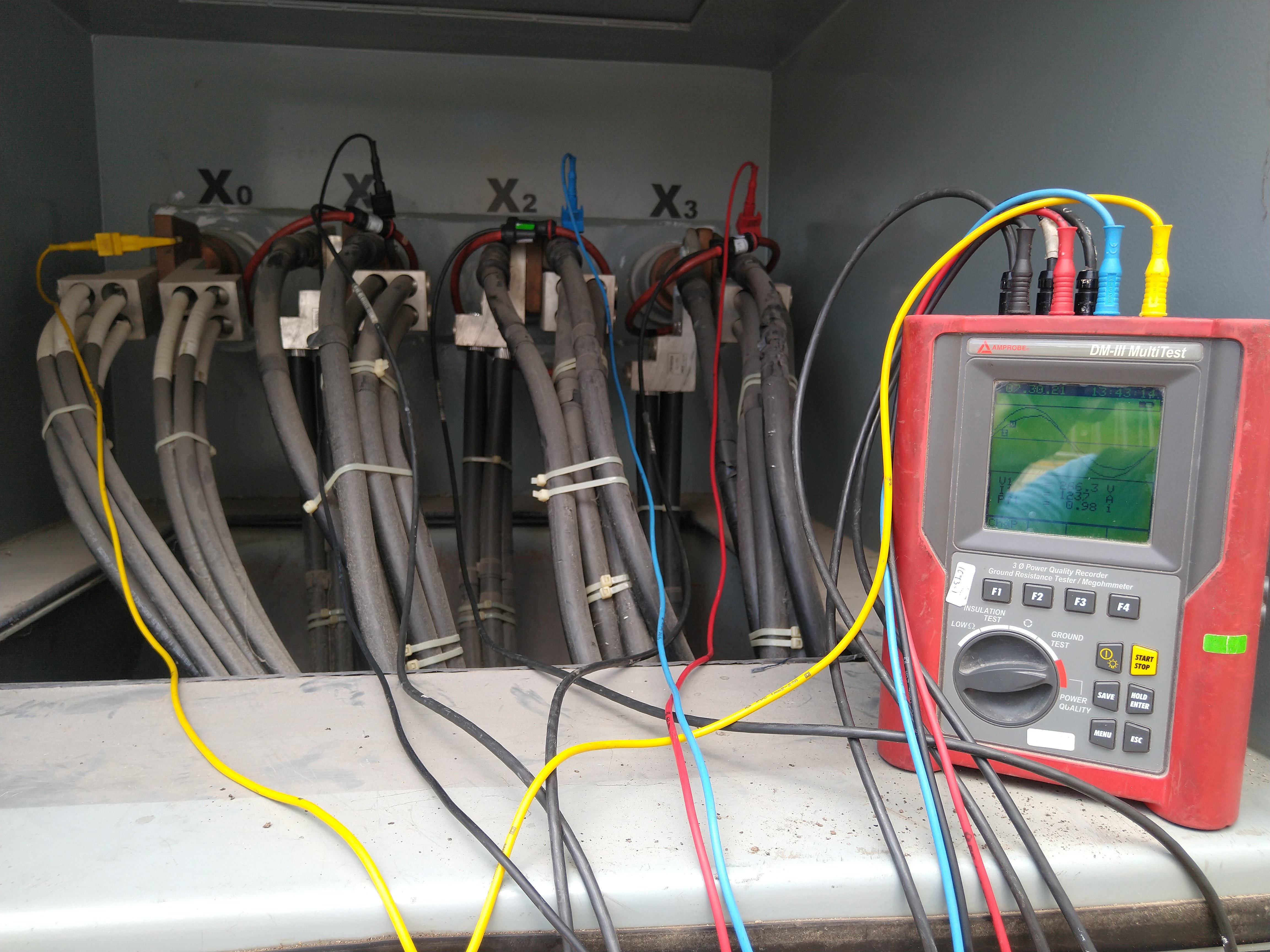 Caja de conexiones eléctricas con cables etiquetados y multímetro digital rojo midiendo parámetros eléctricos.
