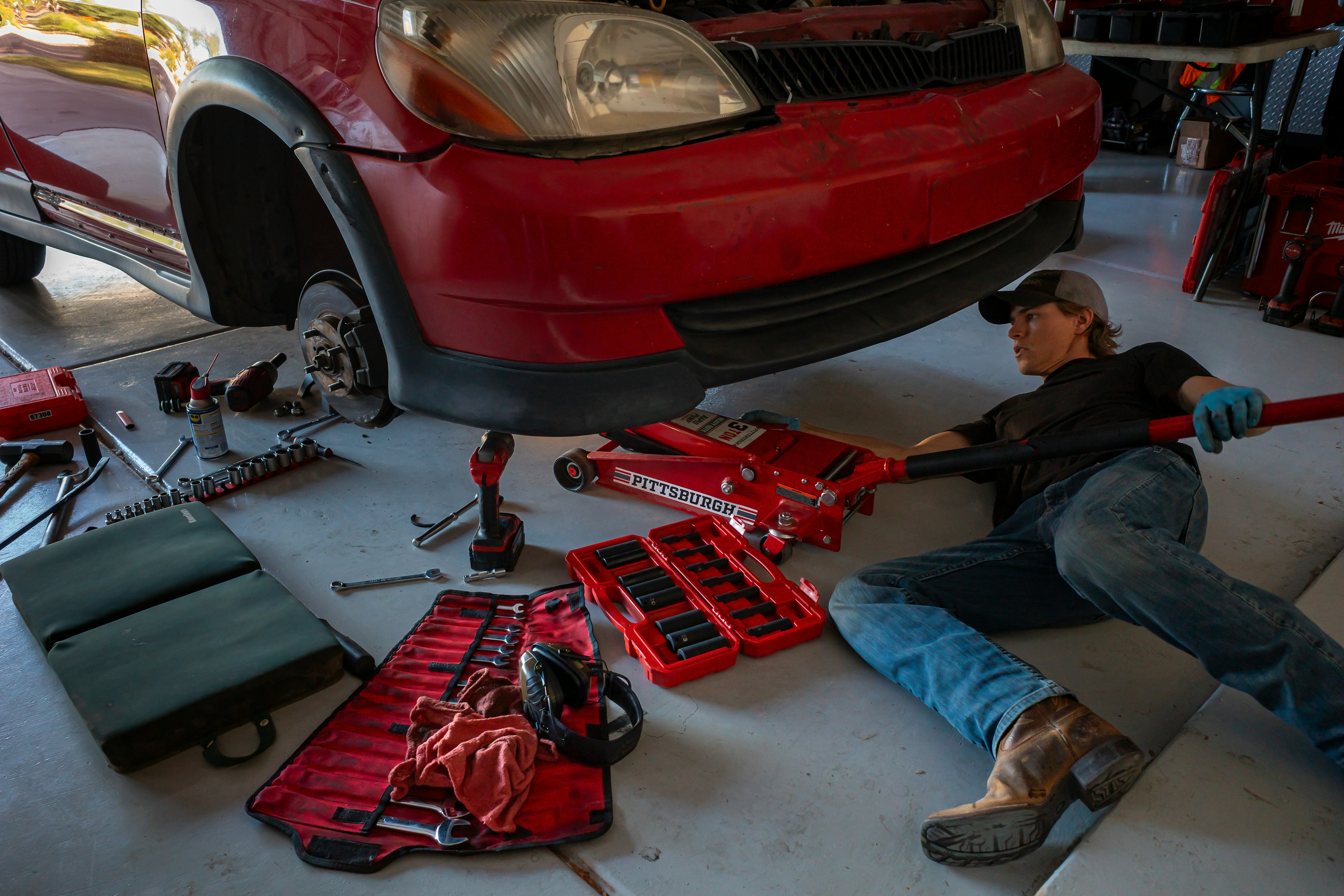 Person working on a red car using a jack in a garage, surrounded by various tools including a wrench set, screwdriver, and drill.