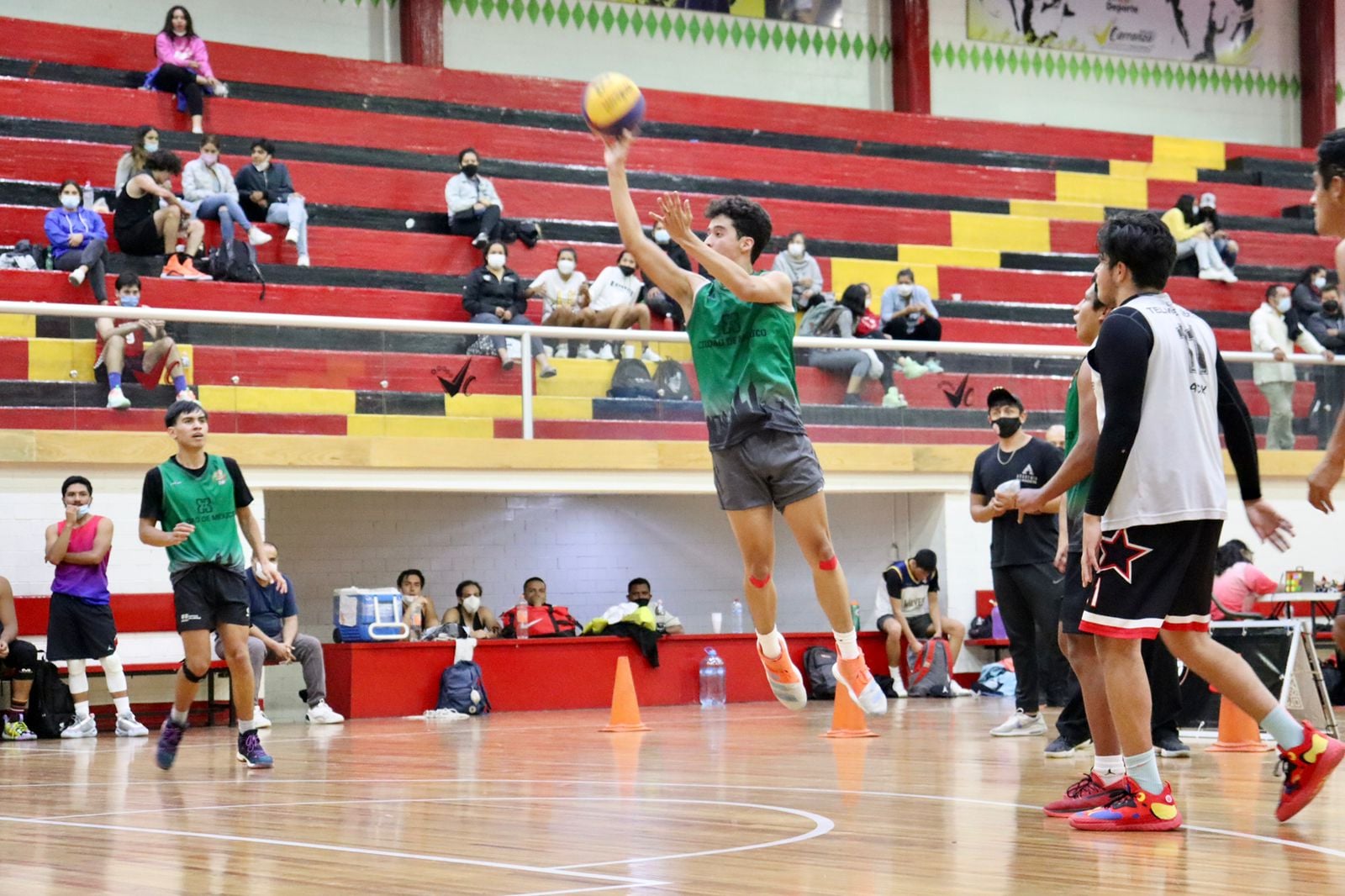 Jugador de baloncesto saltando para lanzar el balón durante un partido en un gimnasio con espectadores en las gradas.