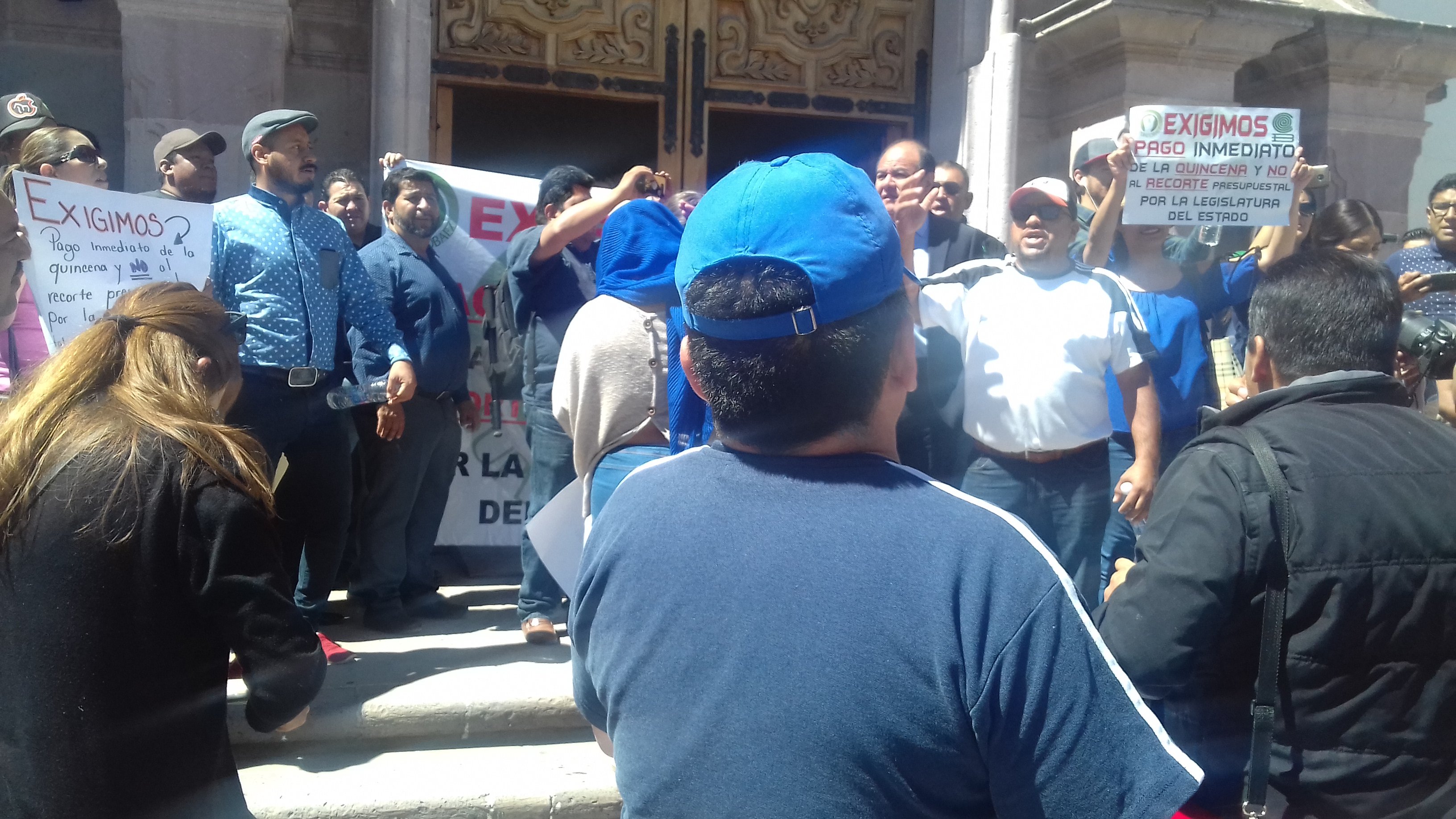 Manifestación en una escalinata con personas sosteniendo pancartas que exigen pago inmediato y protestan contra recortes presupuestarios.