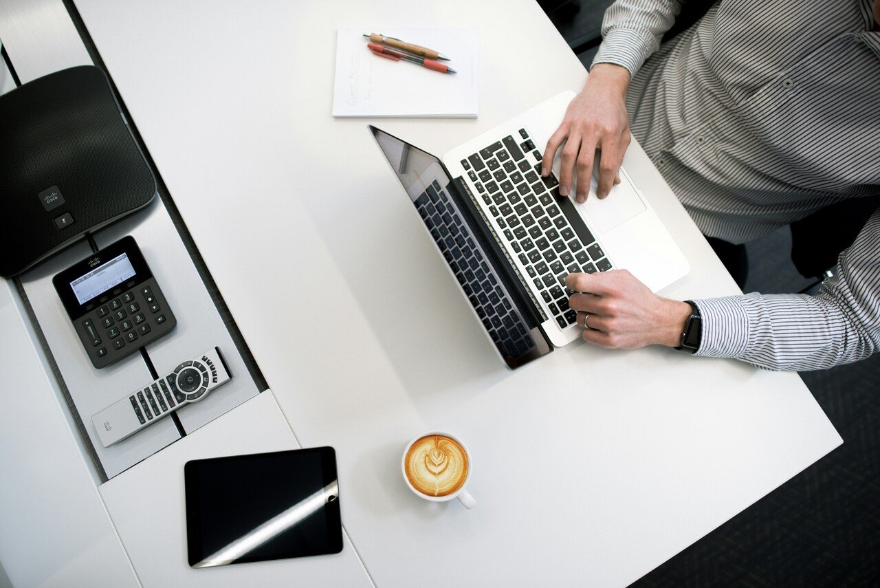 Hombre trabajando en laptop sobre escritorio blanco con cafetera, libreta, teléfono, y tablet.