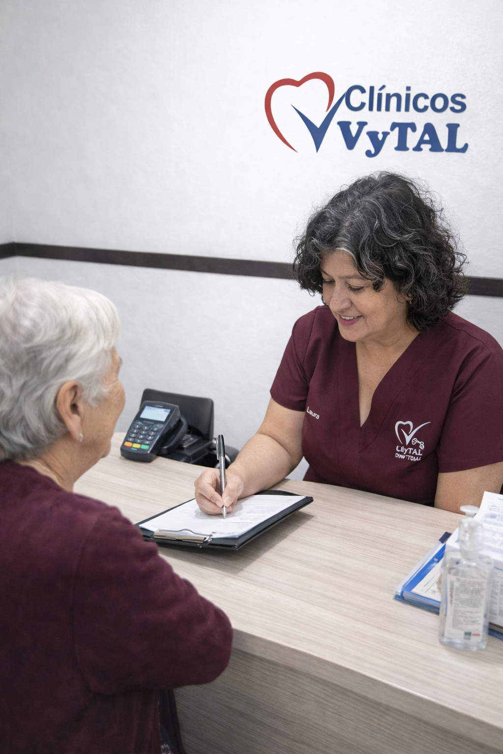 Mujer en uniforme de clínica llenando formularios en una consulta con una paciente en Clínicos VyTAL.