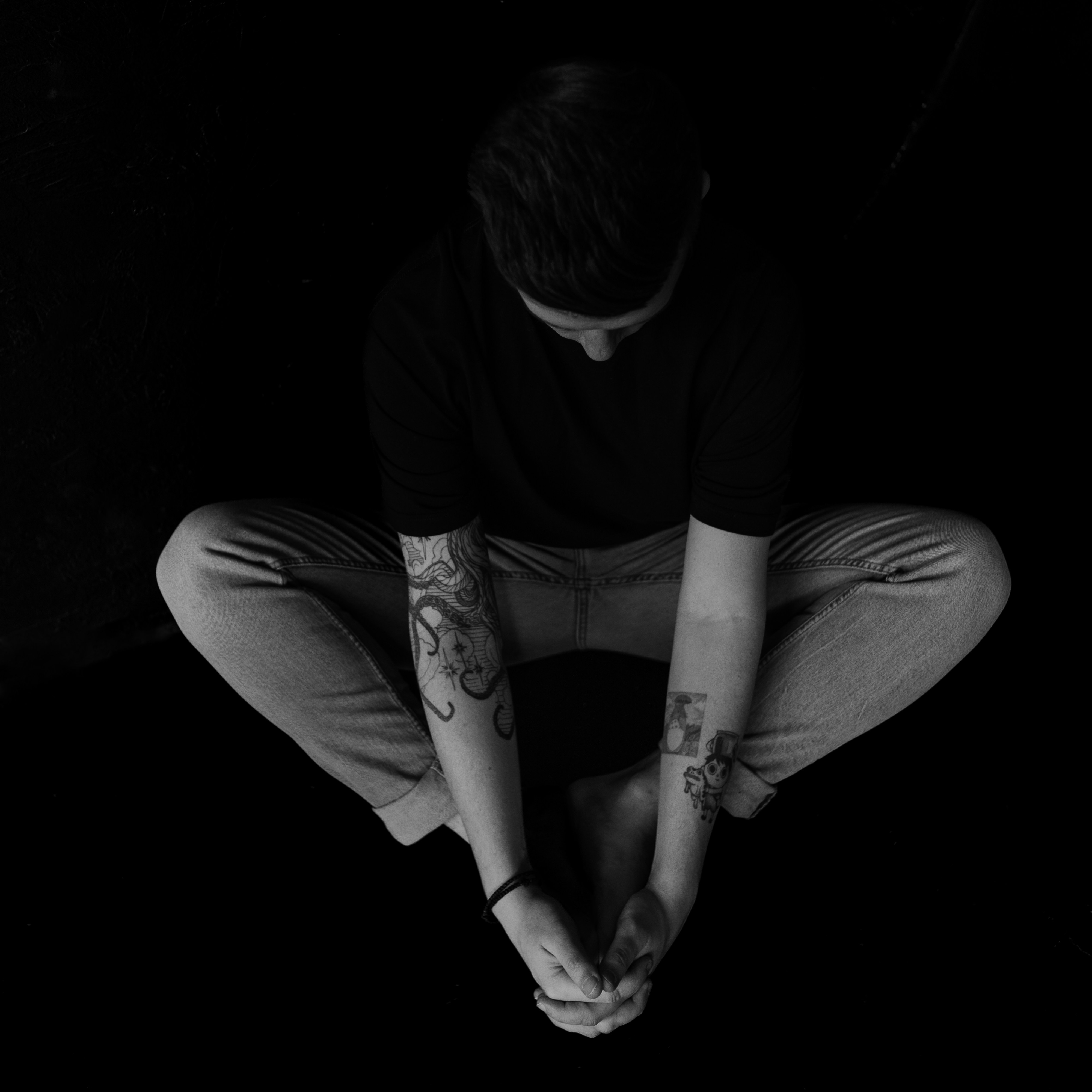 Persona sentada con brazos tatuados en forma de círculo, mirada hacia abajo, en un entorno oscuro blanco y negro.